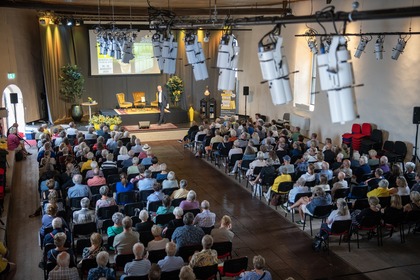 Föreläsning med stor publik i en ljus sal, en person står på scenen framför en projektorduk.