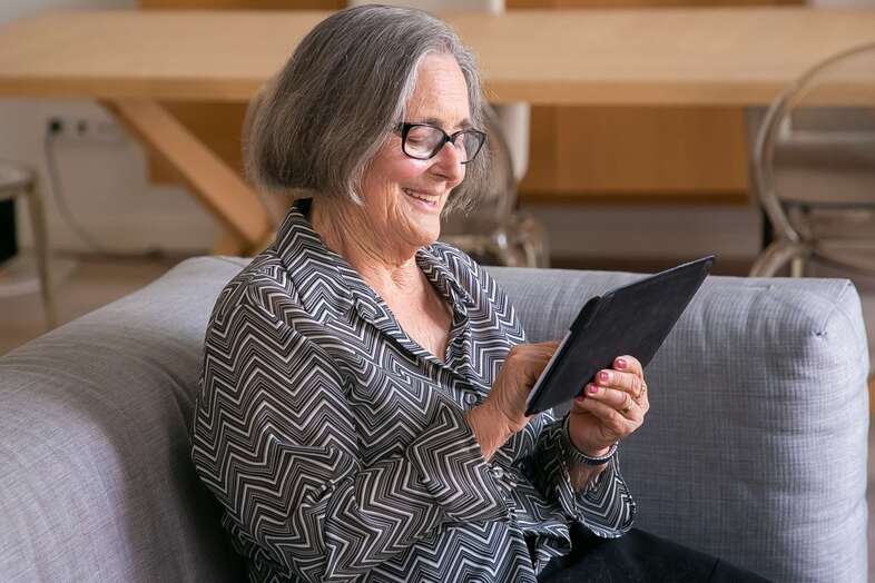 En äldre kvinna sitter i en grå soffa och bläddrar på en iPad.