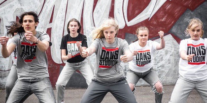 Fem unga personer dansar energiskt framför en graffitivägg, alla bär t-shirts med texten 'Dansstudio'.