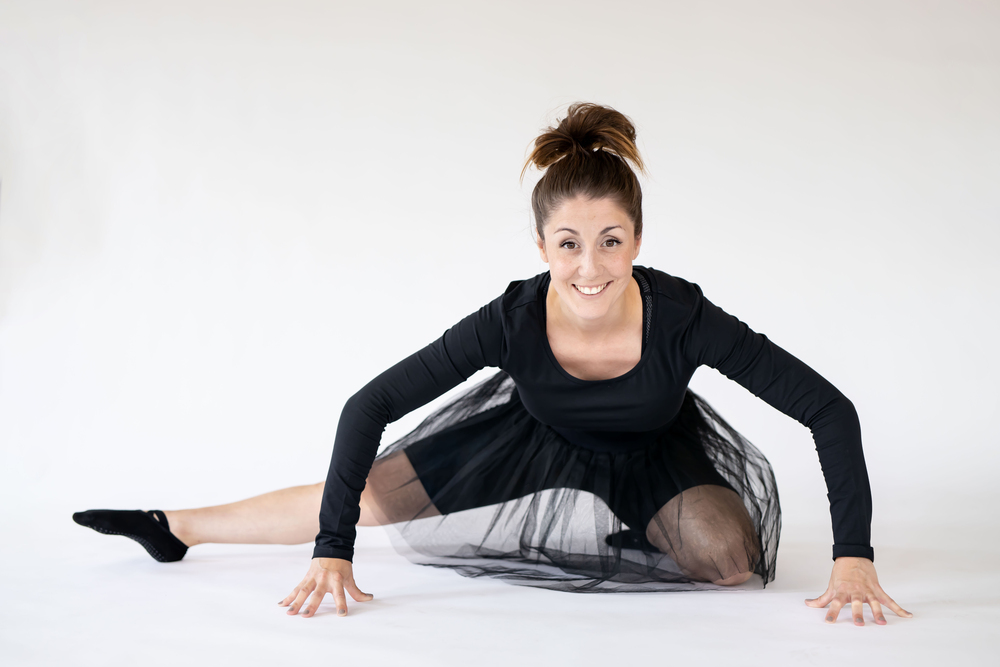 Kvinna i svart klänning gör en dansstretch på golvet och ler mot kameran.