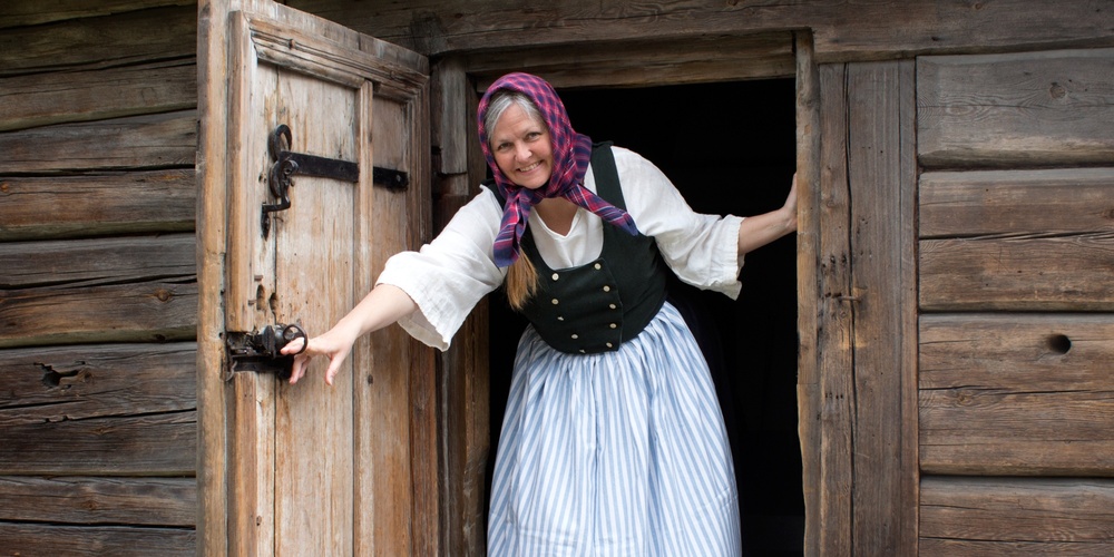 Kvinna i traditionella kläder står i dörröppningen till en gammal timmerstuga och ler.