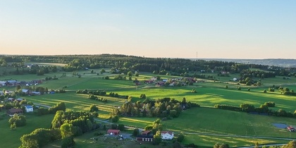 Landskapsvy över gröna fält, skogsdungar och några gårdar under en klar blå himmel.