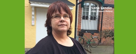 Kvinna med glasögon står utomhus framför gula och röda byggnader, cykel syns i bakgrunden.