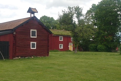 Två röda trähus på en grön gräsmatta med träd och molnig himmel i bakgrunden.