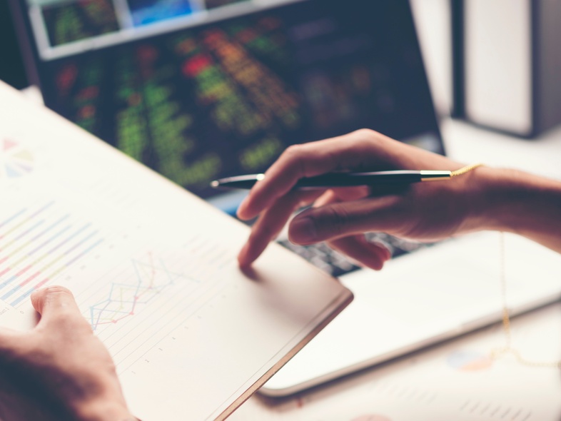 En person granskar diagram och grafer på ett papper med en penna, med en bärbar dator i bakgrunden.