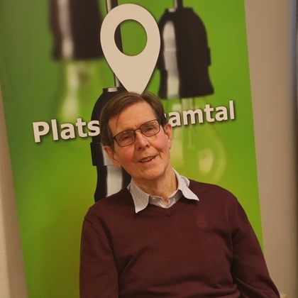 Man med glasögon sitter vid ett bord framför en grön skylt med texten 'Plats för samtal'.