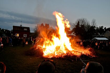 Stor brasa brinner på en gräsmatta omgiven av människor under kvällstid.