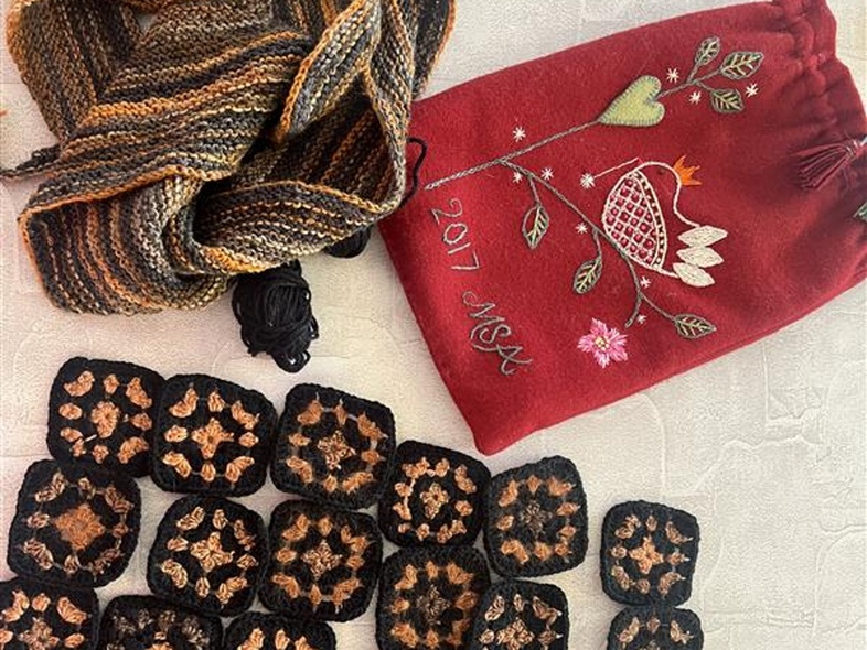 Flera virkade rutor i svart och brunt, en stickad halsduk och en röd påse med broderi på ett bord.