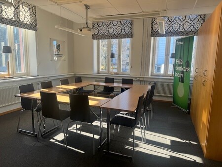 Ljust konferensrum med rektangulärt bord, svarta stolar och stora fönster med mönstrade gardiner.