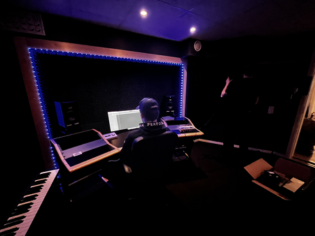 En person sitter vid ett mixerbord i en mörk inspelningsstudio med blå LED-belysning runt arbetsytan.