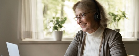 Äldre kvinna med hörlurar deltar i videosamtal vid laptop, antecknar i block vid ett bord.