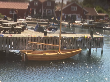 En liten träsegelbåt ligger förtöjd vid en brygga i en småbåtshamn med röda stugor i bakgrunden.