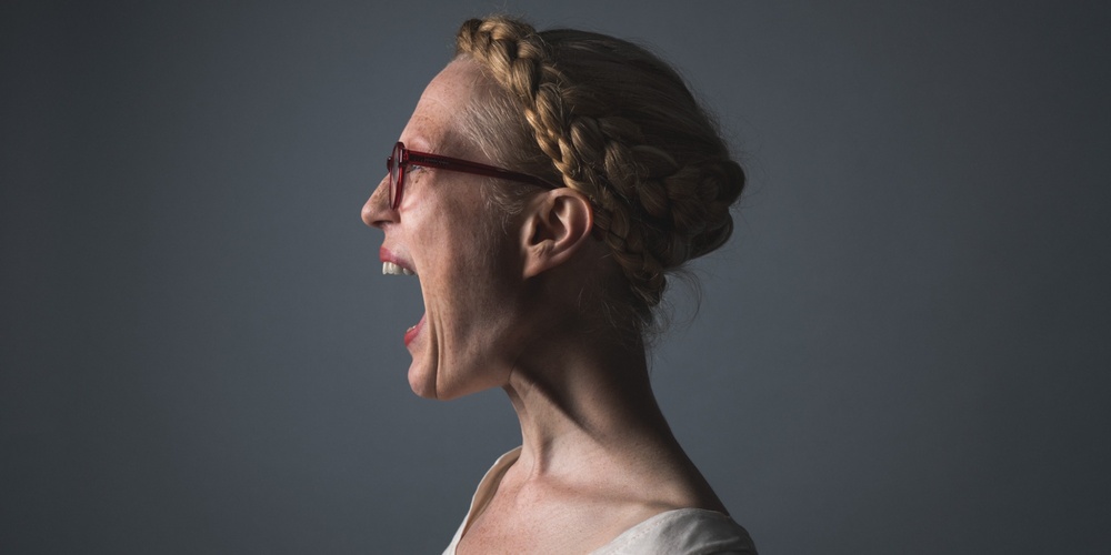 Kvinna med flätat hår och glasögon skriker i profil mot en mörk bakgrund.