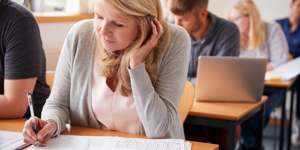 Kvinna sitter i klassrum och skriver på papper, omgiven av andra vuxna studenter.