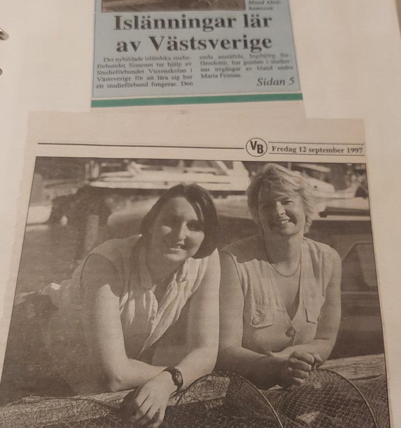 Två kvinnor sitter och ler vid en brygga, med båtar i bakgrunden. Bilden är svartvit och från en tidningsartikel.