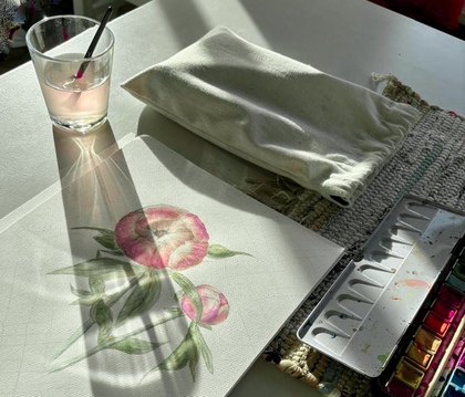 Akvarellmålning av blommor på ett bord med färgpalett, glas med pensel och tygpåse i solljus.