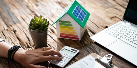 En person använder miniräknare vid ett skrivbord med laptop, pappersarbete, växt och modellhus med energimärkning.