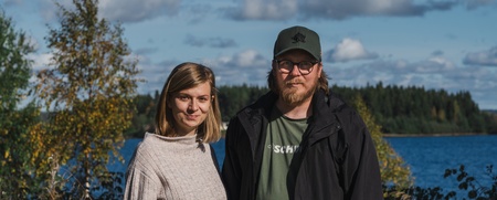 En kvinna och en man står utomhus vid en sjö med skog i bakgrunden under en solig dag.