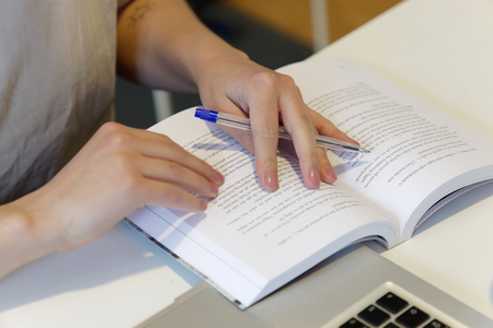 En person läser en bok och markerar text med en penna vid ett skrivbord med en bärbar dator.