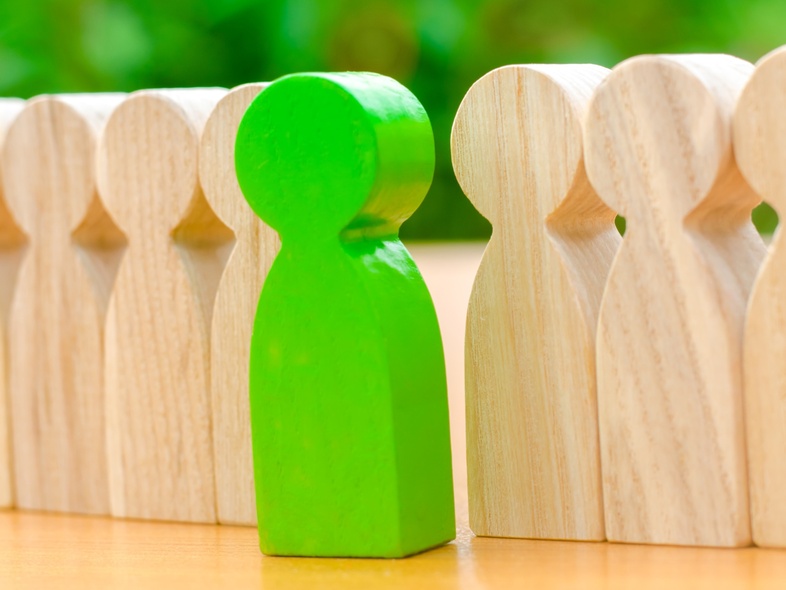 En grön träfigur står mellan två rader av ljusa träfigurer på ett bord med grön bakgrund.