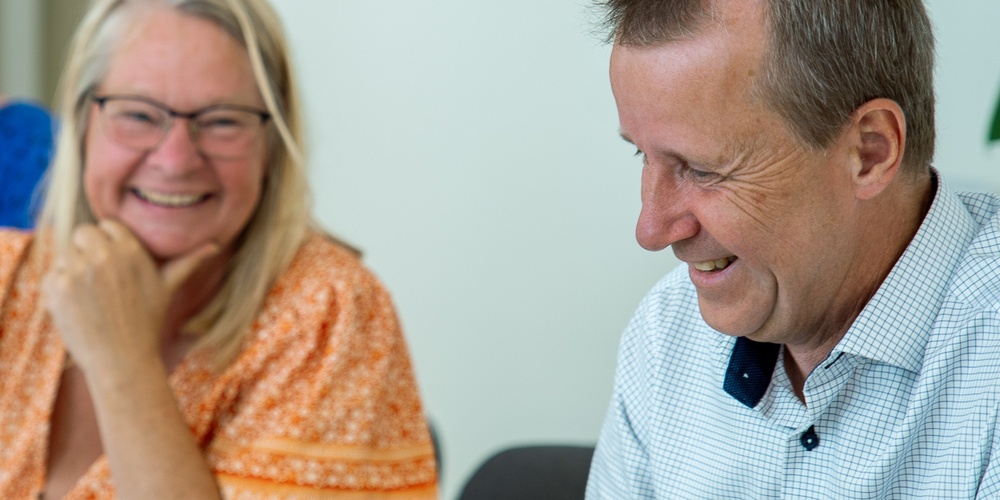 Två glada personer sitter vid ett bord och samtalar i ett ljust kontorsrum.