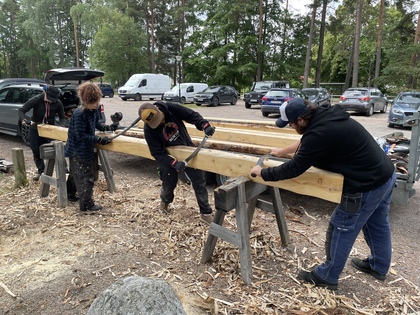 Fyra personer bearbetar stora träbalkar utomhus med handverktyg, omgivna av bilar och träd.