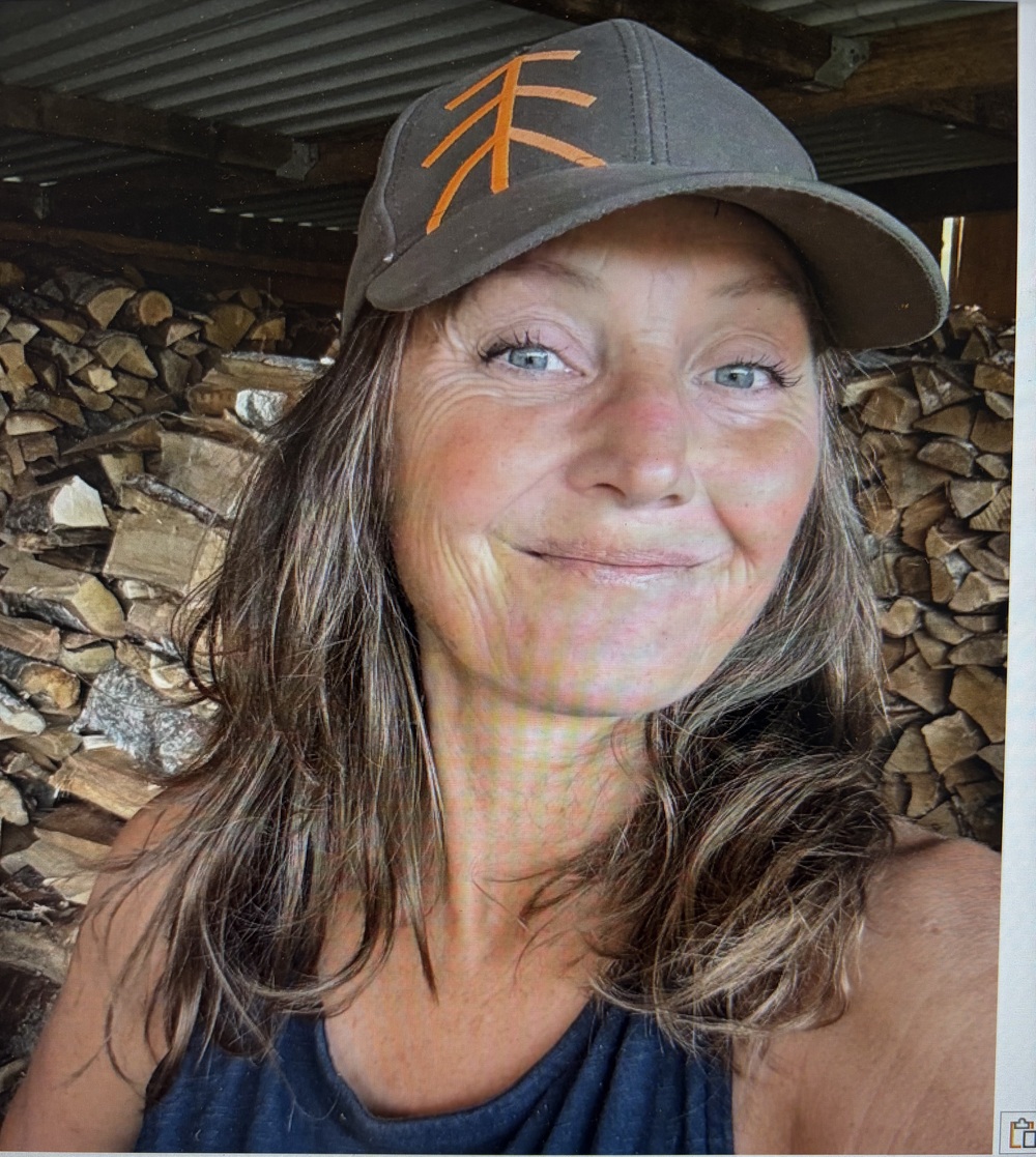 Kvinna med långt hår och keps tar en selfie framför en stapel ved inomhus.