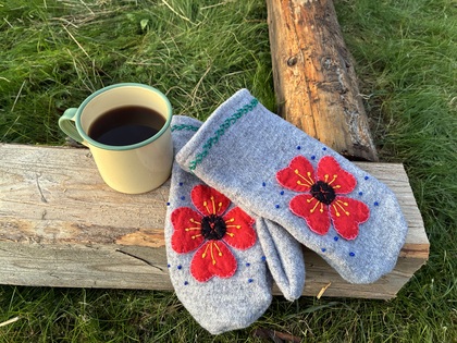 En kopp svart kaffe och ett par grå vantar med röda blommor ligger på en träbit utomhus på gräs.