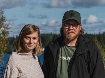 En kvinna och en man står utomhus vid en sjö med skog i bakgrunden under en solig dag.
