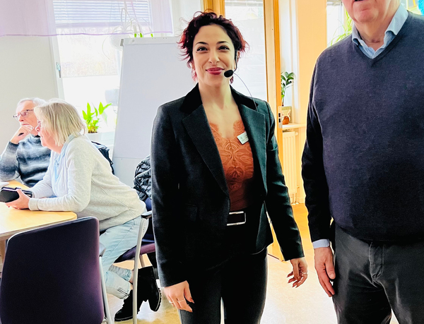 Kvinna med headset står och ler i ett ljust rum, flera personer sitter vid ett bord i bakgrunden.