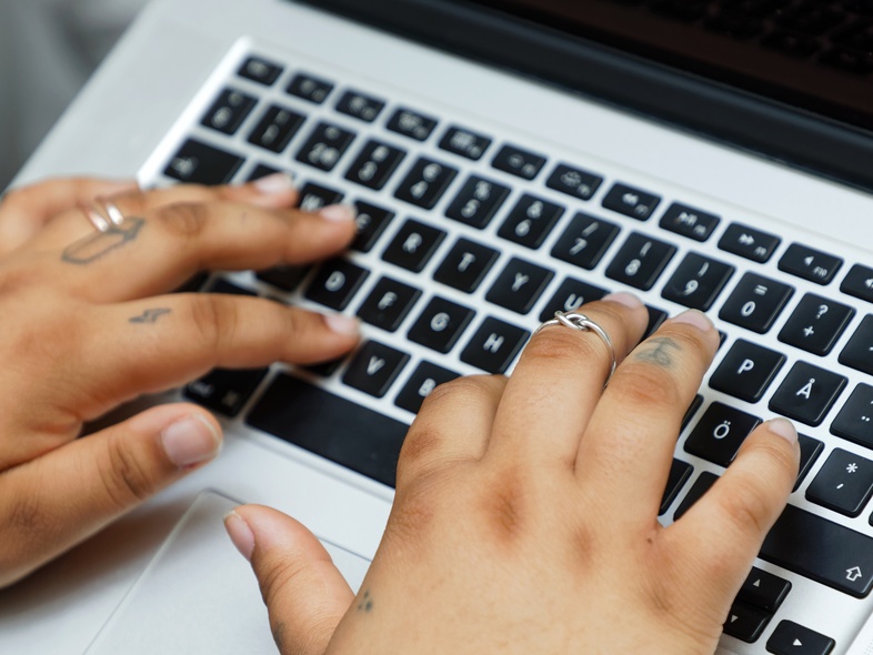Två händer med ringar och tatueringar skriver på ett svartvitt tangentbord på en bärbar dator.