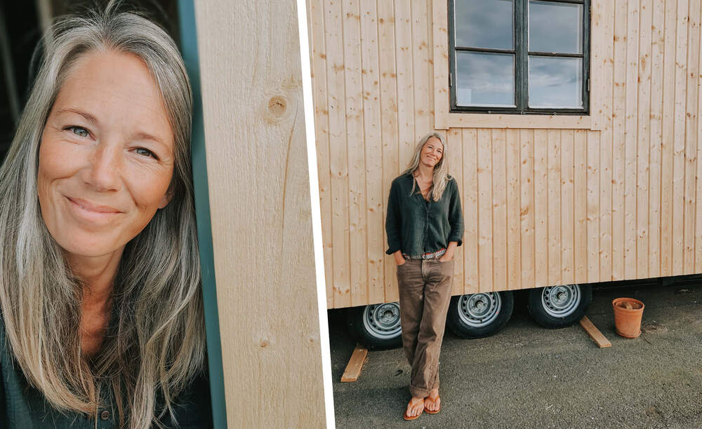Person med långt grått hår står framför en träbyggnad med hjul och en kruka på marken.