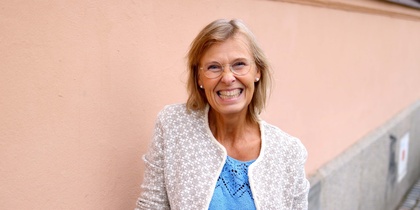 Leende äldre kvinna med glasögon står mot en ljusrosa vägg utomhus.