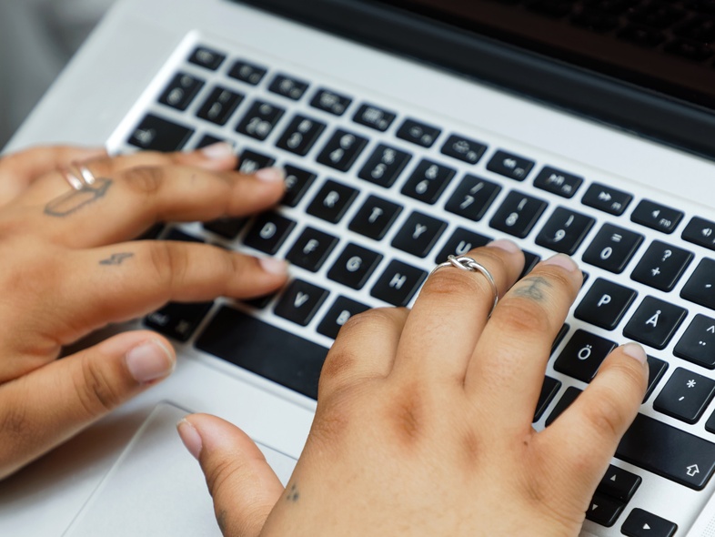 Två händer med ringar och tatueringar skriver på ett svartvitt tangentbord på en bärbar dator.