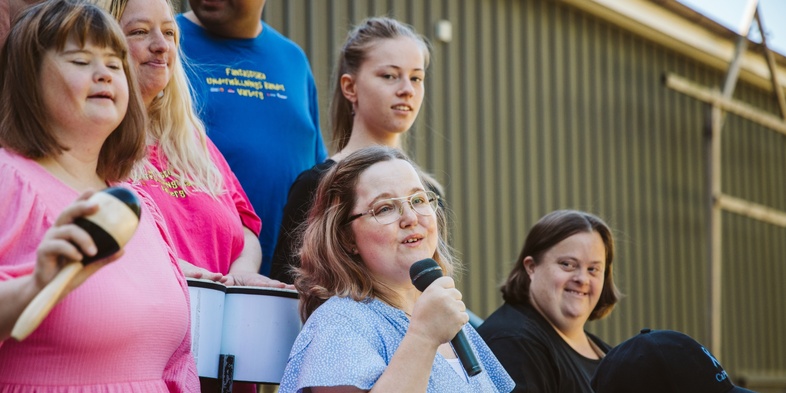En grupp människor, flera med mikrofon och musikinstrument, står utomhus och sjunger tillsammans.