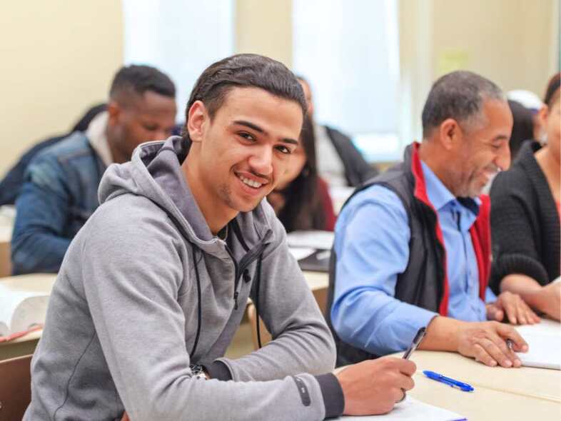 En grupp personer sitter i ett klassrum och arbetar. En person är vänd mot kameran och ler.