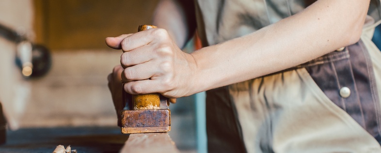 En person i arbetskläder hyvlar trä i en snickarverkstad, fokus på händer och verktyg.