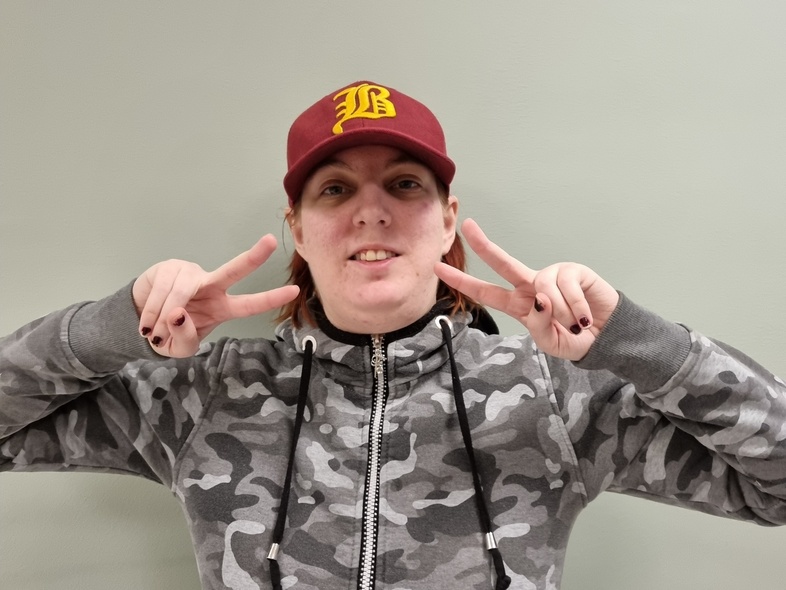 Person i grå kamouflagehoodie och röd keps gör peace-tecken med båda händer mot en ljus vägg.