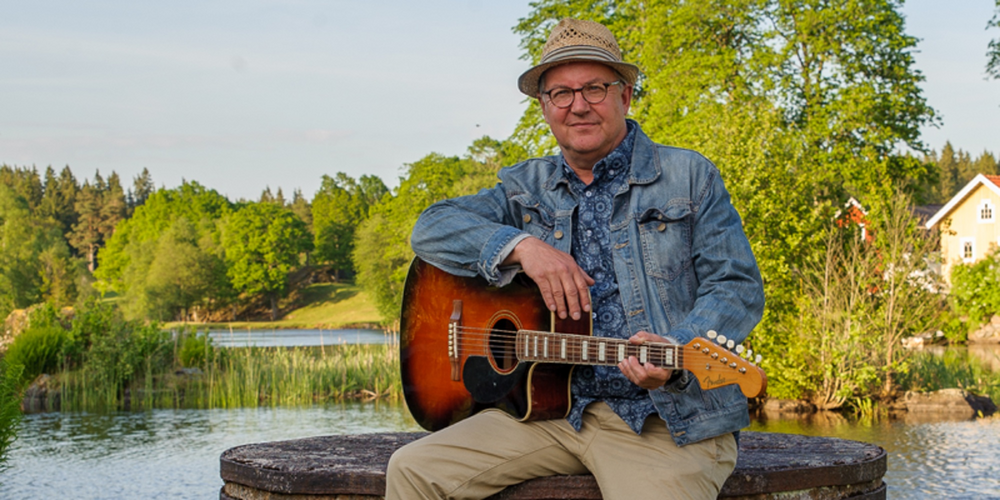 En person sitter utomhus på en rund bänk och spelar akustisk gitarr vid en sjö.