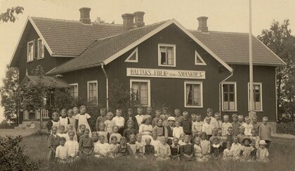 Gammalt svartvitt foto av en stor grupp barn och några vuxna framför en skolbyggnad med skylten 'Baltaks folk- och småskola'.