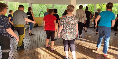 En grupp vuxna dansar tillsammans på ett trägolv i en ljus paviljong med stora fönster.