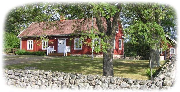 Röd trävilla med vita knutar, omgiven av träd och en stenmur i lantlig miljö.