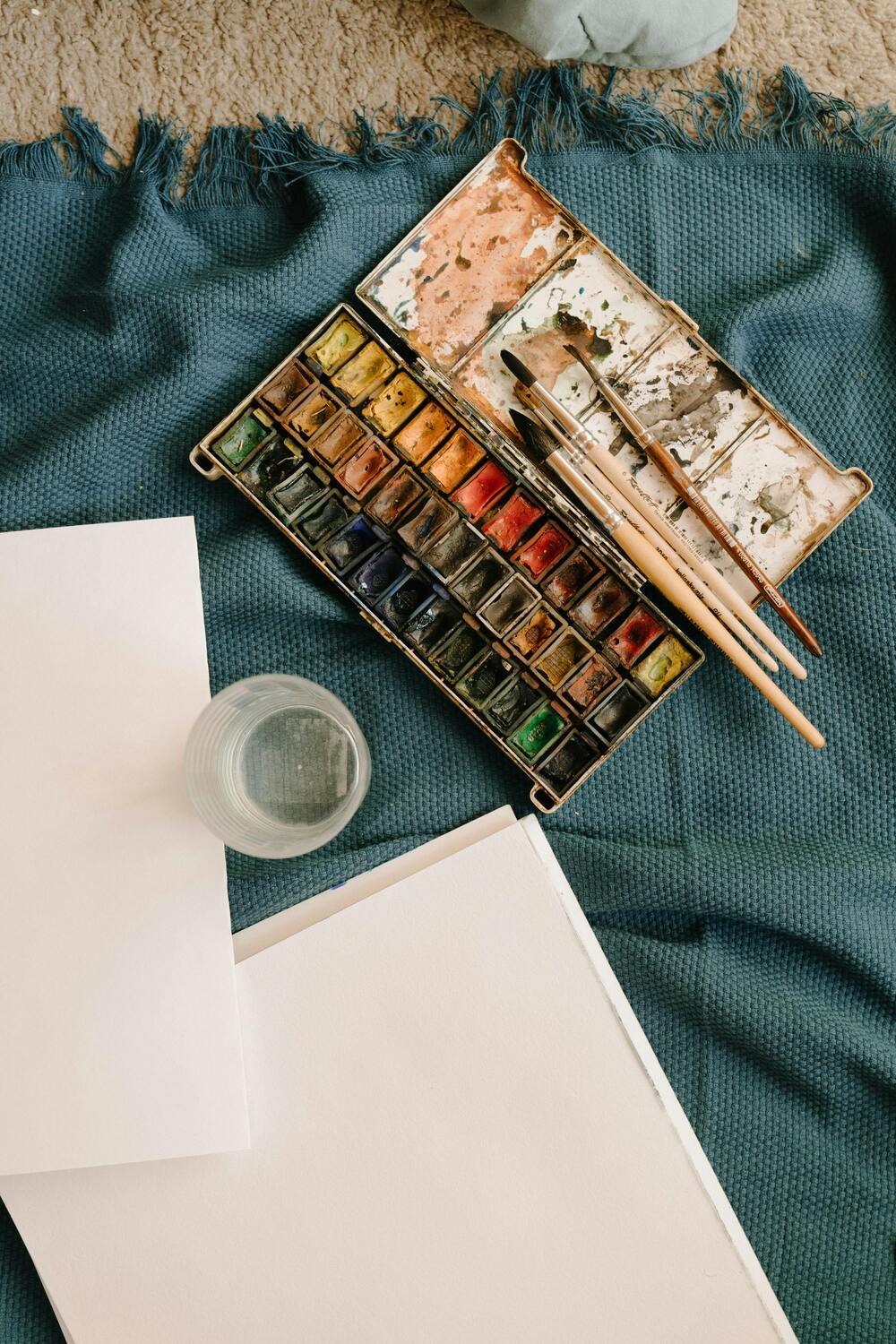 Akvarellfärger, penslar, papper och ett glas vatten ligger på en blå filt, redo för målning.