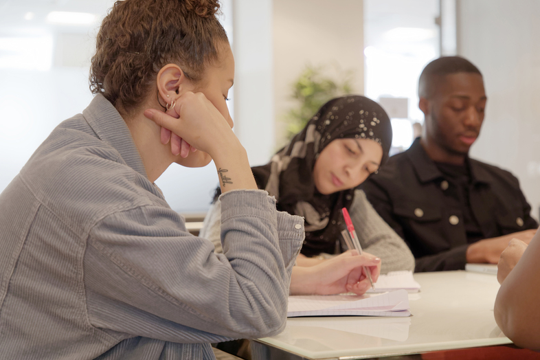 Tre unga personer sitter vid ett bord och studerar tillsammans, en skriver i ett block.