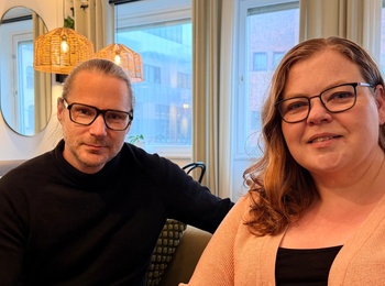 Två personer med glasögon sitter i en modern, mysig kafémiljö med stora fönster och varma lampor.