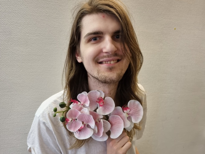 Person med långt hår i vit t-shirt håller en bukett rosa orkidéer framför bröstet, ansiktet är suddat.