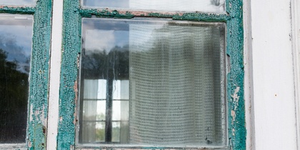 Närbild av ett gammalt fönster med avskavd grön färg och gardin på insidan.