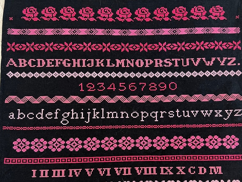 Broderad duk med alfabetet, siffror och mönster i rosa och rött på svart bakgrund.