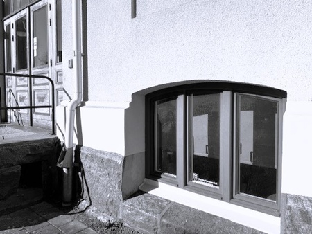 En källarfönster i en vit putsad byggnad bredvid en entré med trappa och räcke.