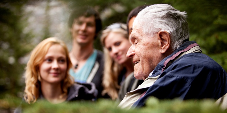 En äldre man pratar med en grupp yngre vuxna utomhus i en grönskande skogsmiljö.